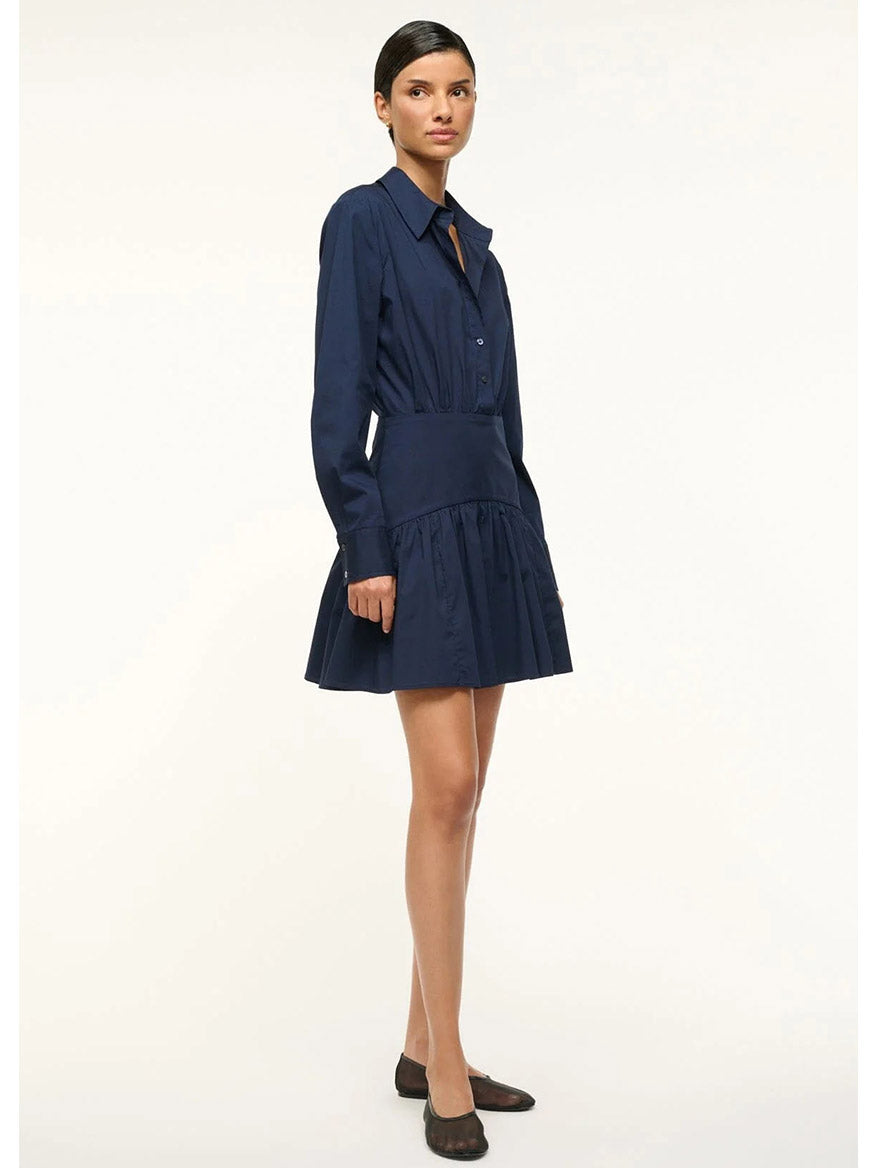 A person stands against a plain background, wearing a chic STAUD Rocco Mini Dress in Navy paired with black flats.