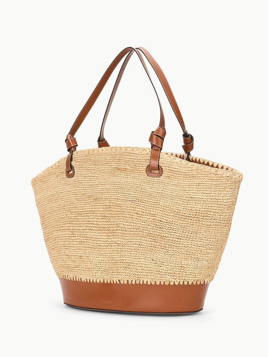 The STAUD Squillo Raffia Tote in Natural/Tan is a spacious tote bag made from raffia, with brown leather handles and trim, showcasing stitched details and a minimalist design.