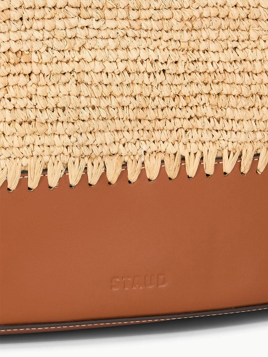 A detailed view of the STAUD Squillo Raffia Tote in Natural/Tan, featuring brown leather handles, visible stitching, and embossed "STAUD" text.