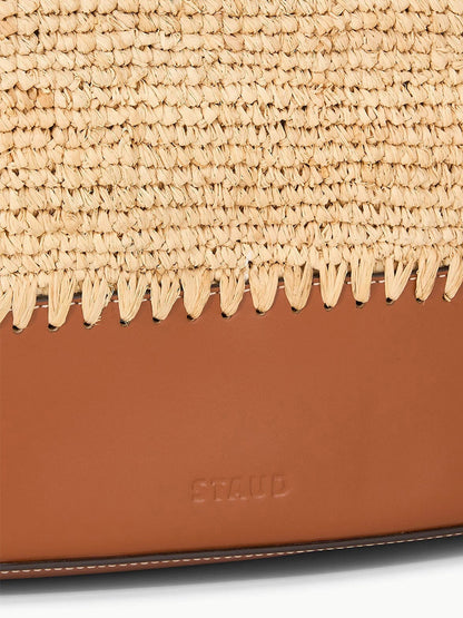 A detailed view of the STAUD Squillo Raffia Tote in Natural/Tan, featuring brown leather handles, visible stitching, and embossed "STAUD" text.