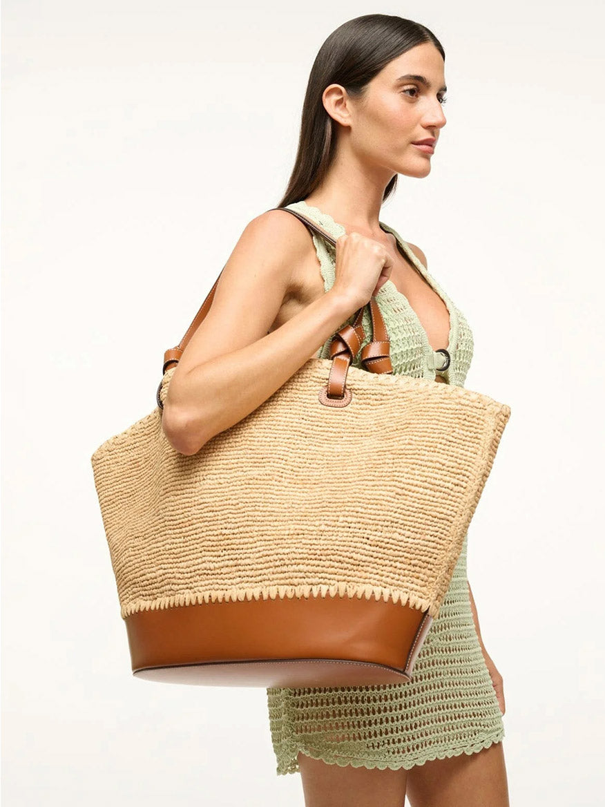 A person in a light green dress holds the STAUD Squillo Raffia Tote in Natural/Tan, featuring stylish leather accents and raffia details, on their shoulder.