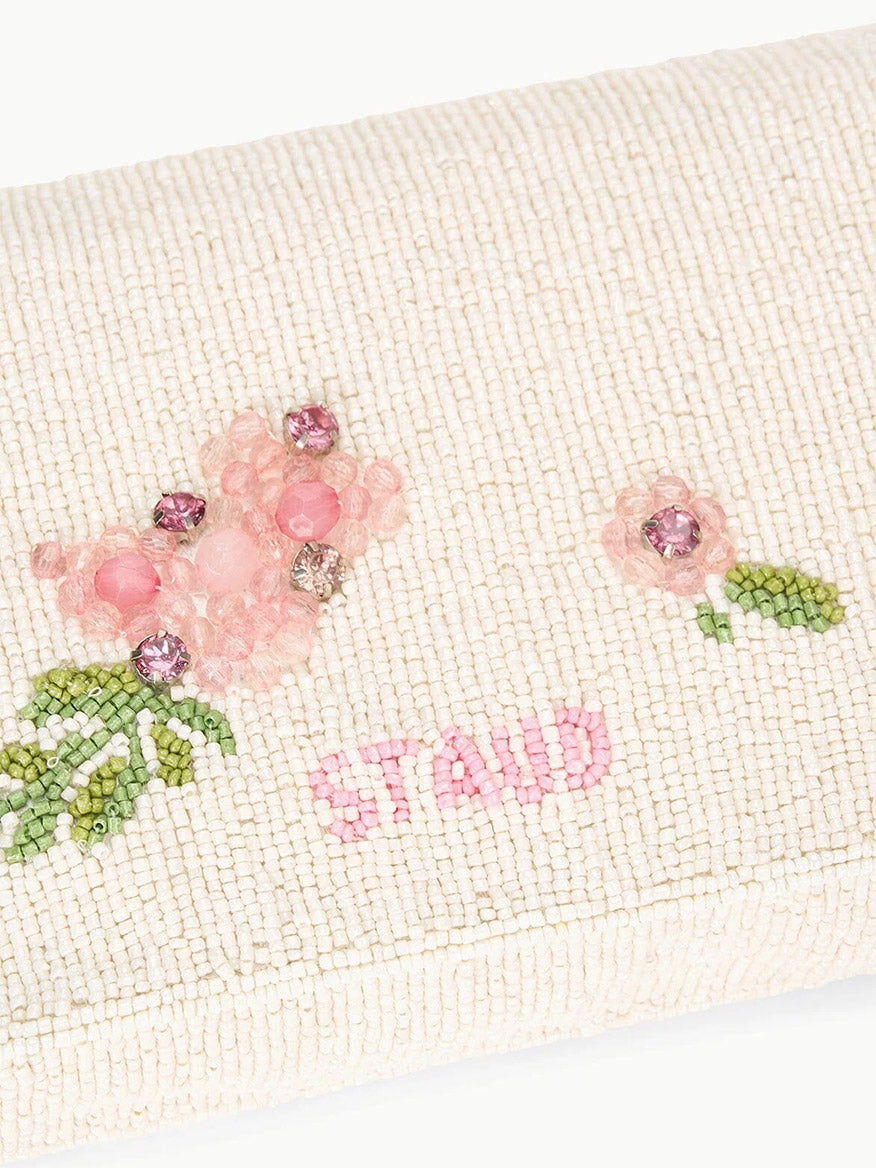 The STAUD Timmy Shoulder Bag in Magnolia is a white clutch adorned with pink beaded flowers and colorful beads, showcasing the word "STAND" in pink beads. This chic accessory can also be used as a beaded vegan leather shoulder bag with a removable strap, providing practicality with its secure magnetic closure.