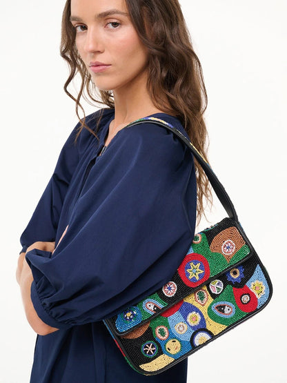A person wearing a navy blue top holds the STAUD Tommy Beaded Bag in Black Millefiori, featuring colorful embroidered circular patterns, over their shoulder.