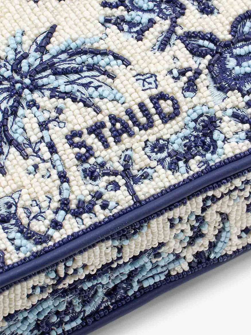 A detailed look at the STAUD Tommy Beaded Bag in Toile de Pup, showcasing hand-beaded artwork with navy blue embroidery spelling "STAUD" amidst intricate palm tree patterns in various shades of blue.