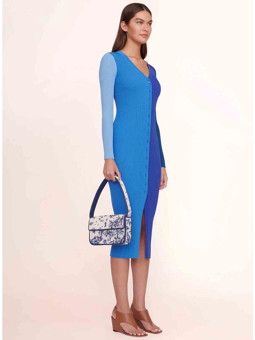 A woman in a blue gradient knit dress holds a STAUD Tommy Beaded Bag in Toile de Pup, standing gracefully against a neutral background.