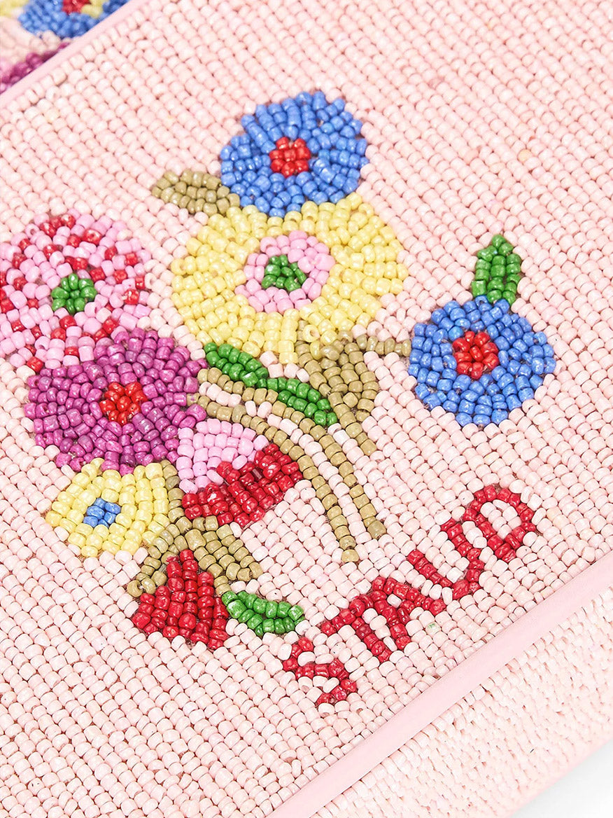 Close-up of a pink beaded fabric featuring 3D floral patterns in red, blue, yellow, and green. The product name "STAUD Tommy Beaded Bag in Wildflowers" is intricately hand-beaded in red at the bottom.