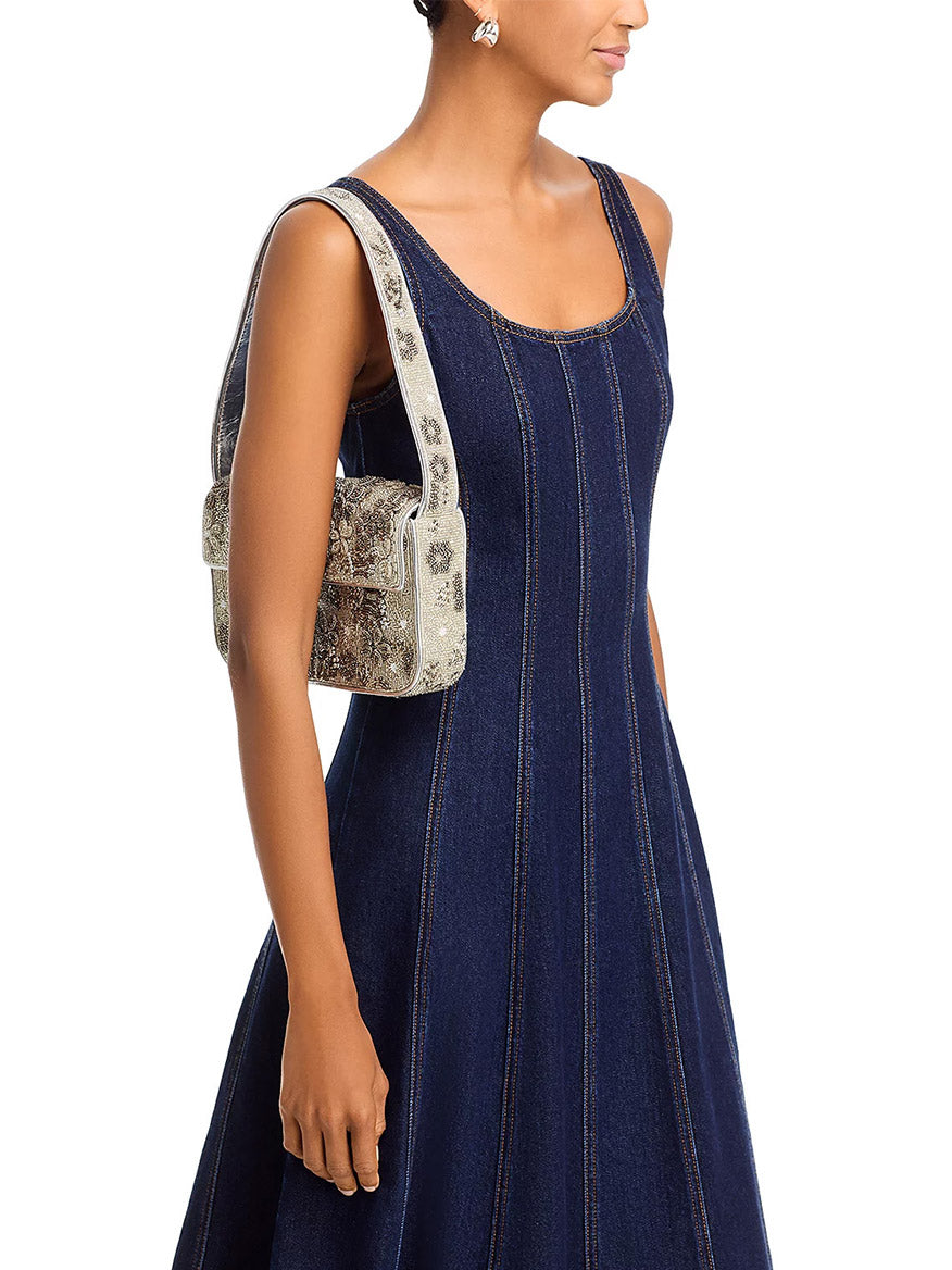 A person in a sleeveless denim dress carries the STAUD Tommy Beaded Bag in Wildflowers on their right shoulder, which is adorned with intricate, embellished 3D flowers.