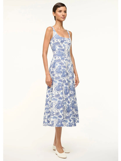 A woman stands against a plain background in the STAUD Wells Sleeveless Dress in Blue Toile, featuring a fitted shape and floral design, paired with white shoes.