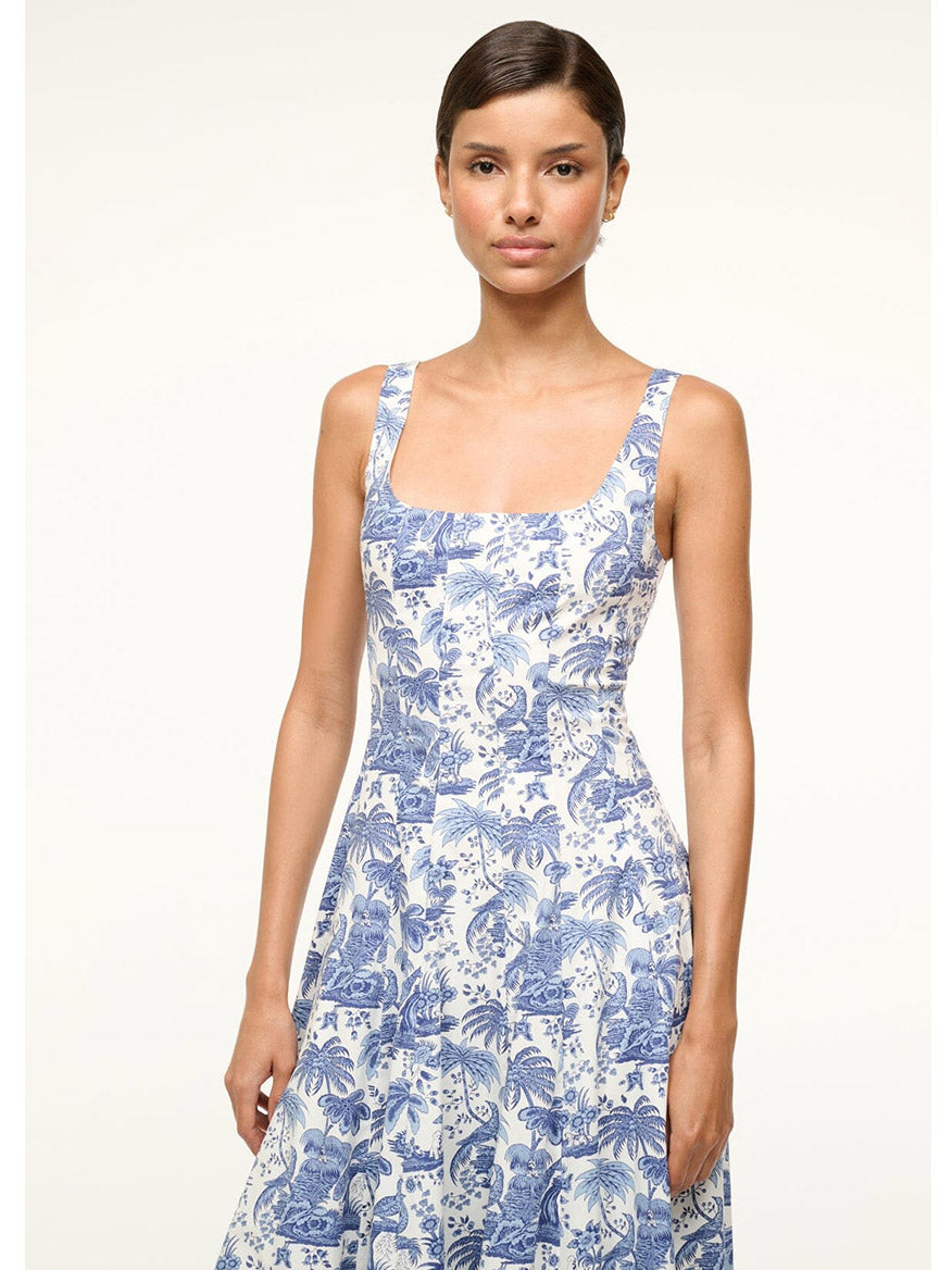 A woman elegantly poses in a STAUD Wells Sleeveless Dress, featuring a fitted shape and blue floral pattern, set against a plain backdrop. The cotton poplin fabric subtly enhances its sophisticated charm and timeless appeal.