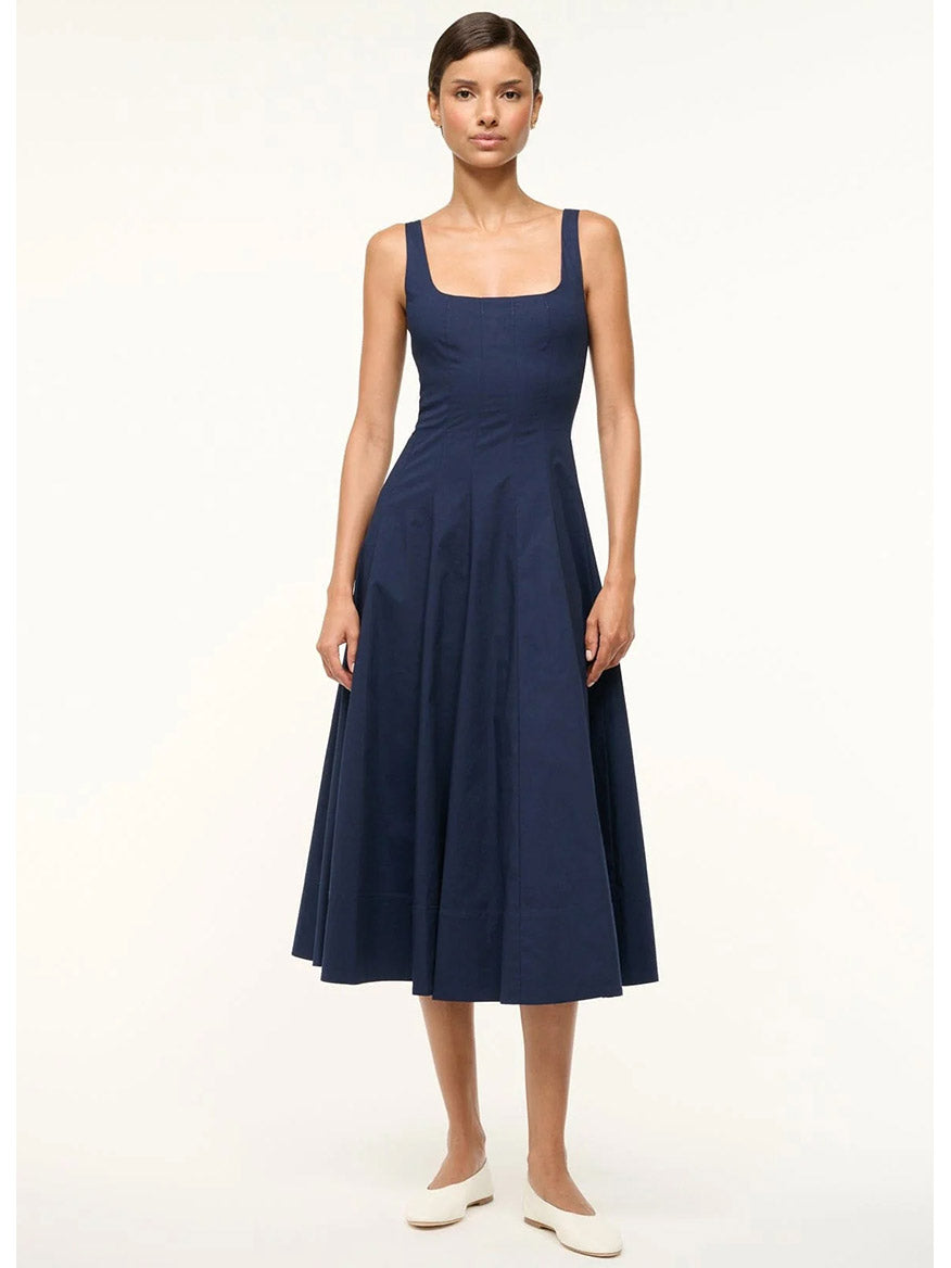 A person wears the STAUD Wells Sleeveless Dress in navy, featuring a corset-style bodice and midi-length skirt, paired with white shoes against a plain white background.