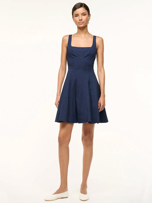 A woman stands against a plain background, wearing the STAUD Mini Wells Dress in Navy with a corset-style square neckline, flared skirt, and white flats.