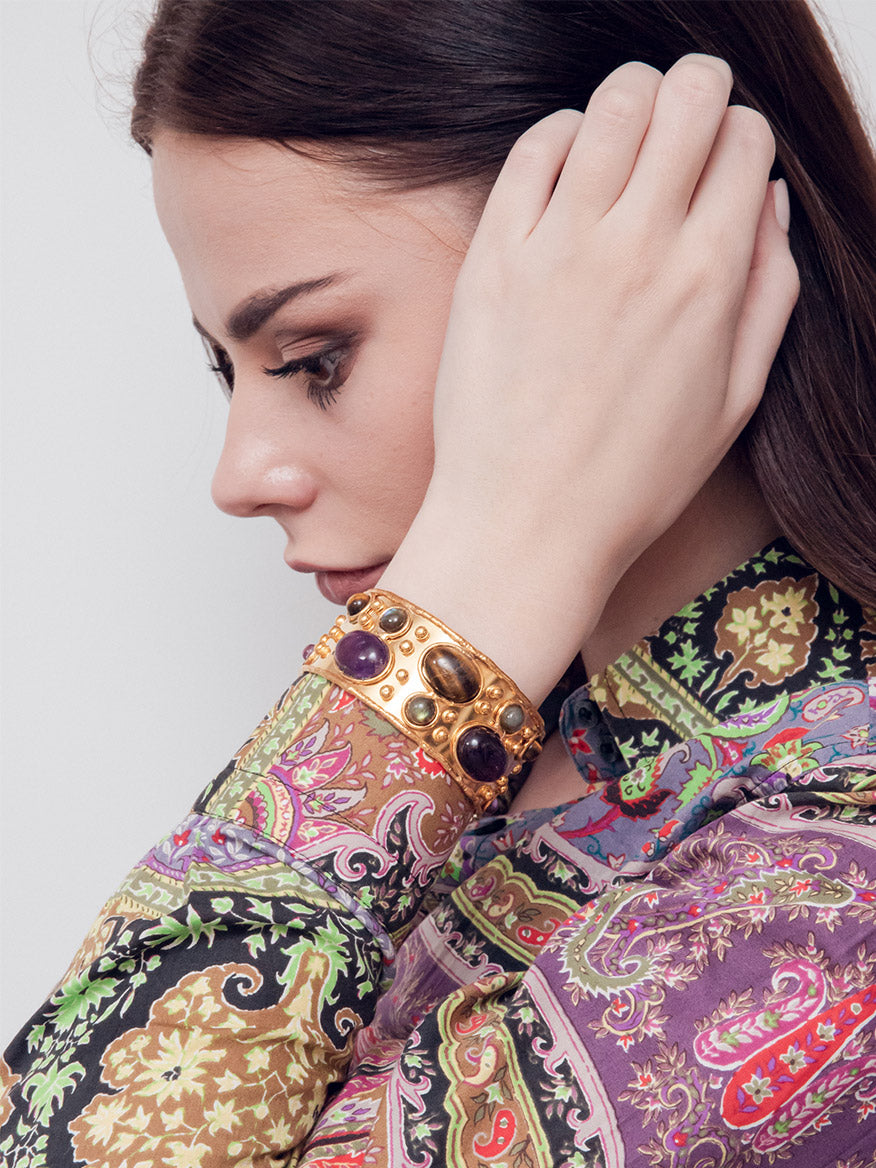 A person with long hair touches their head, wearing a colorful paisley-patterned shirt and the Sylvia Toledano Cuff Byzantine in Amethyst, Tiger Eye, Labradorite & Gold.