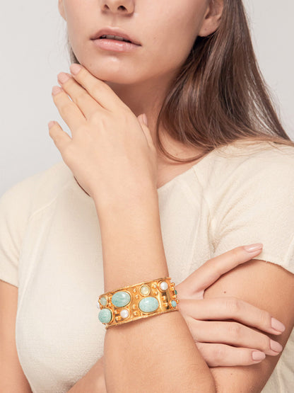 A person wearing a white top poses with their hand near their chin, showcasing the dazzling gold Sylvia Toledano Byzantine cuff bracelet adorned with Amazonite, Pearl, and Pale Green Onyx stones.