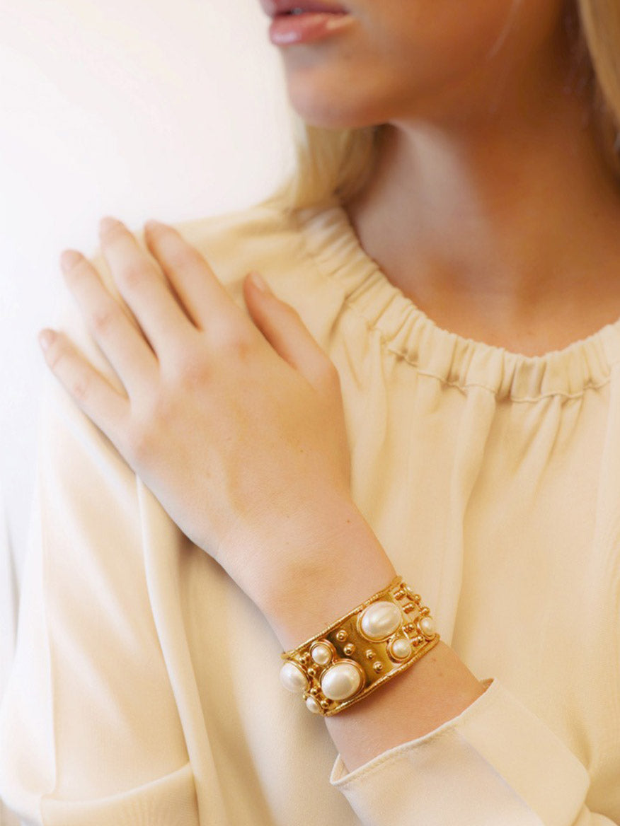 A person wearing a cream blouse and the Sylvia Toledano Cuff Byzantine in Golden Pearl, featuring intricate brass details and adorned with large pearls, on their wrist.