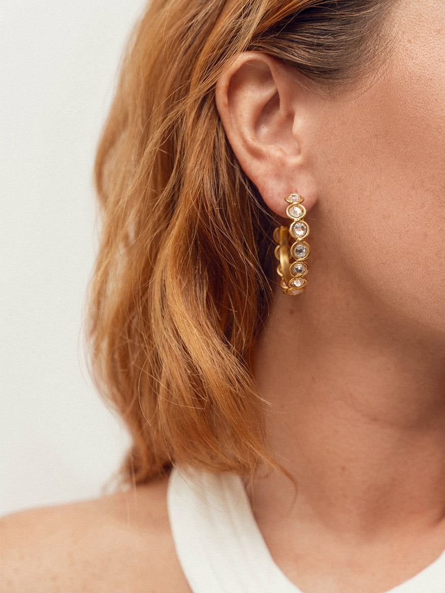 A close-up of a woman's ear wearing fine gold Sylvia Toledano Earrings Candies in Gold & Swarovski Crystals adorned with multiple small gemstones. The woman has light brown hair and is wearing a white sleeveless top.