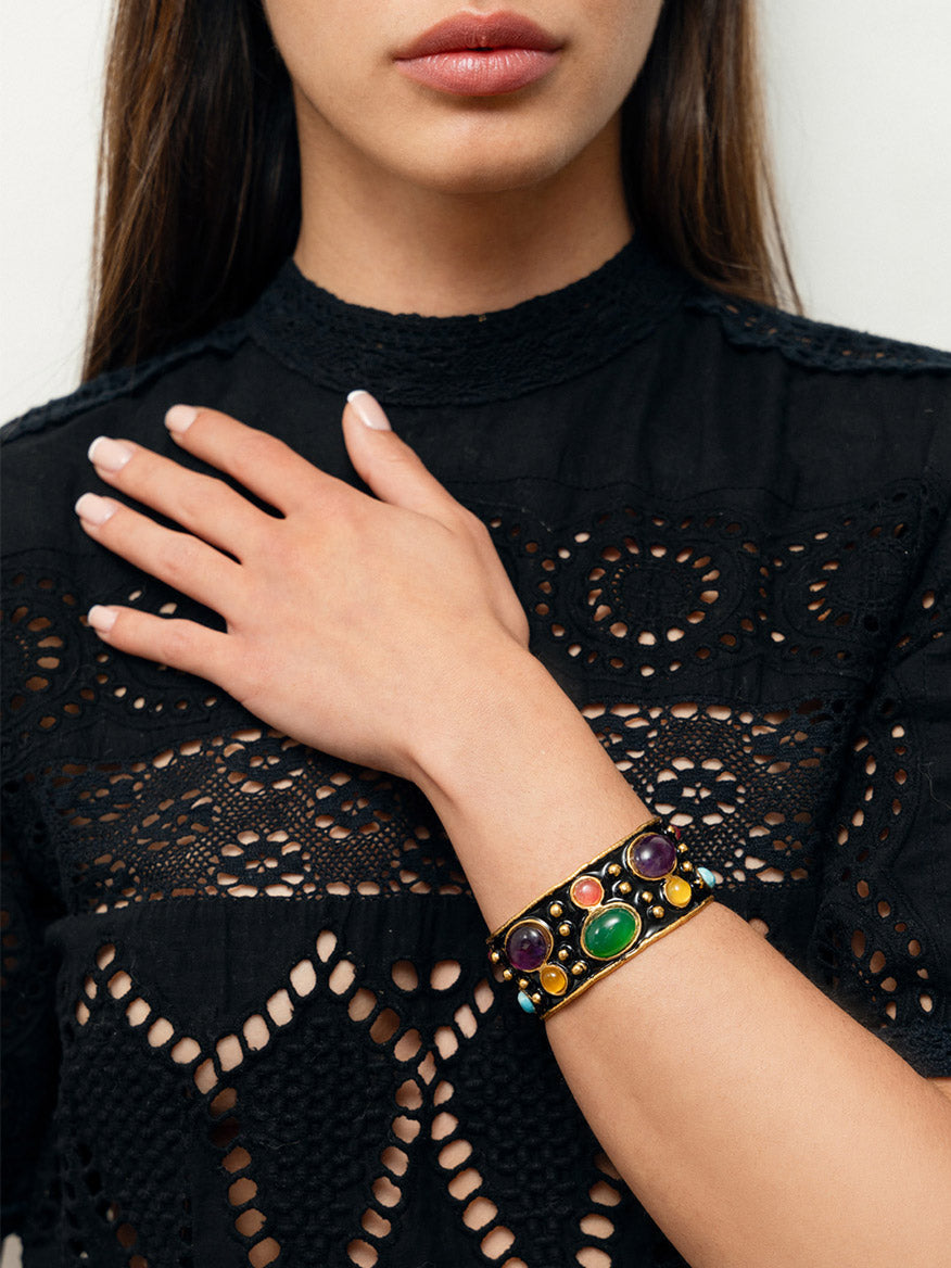 A woman wearing a black lace top accessorized with the Sylvia Toledano Cuff Byzantine in Multi Winter & Black Enamel on her left wrist, featuring colorful gemstones with an enchanting green onyx.