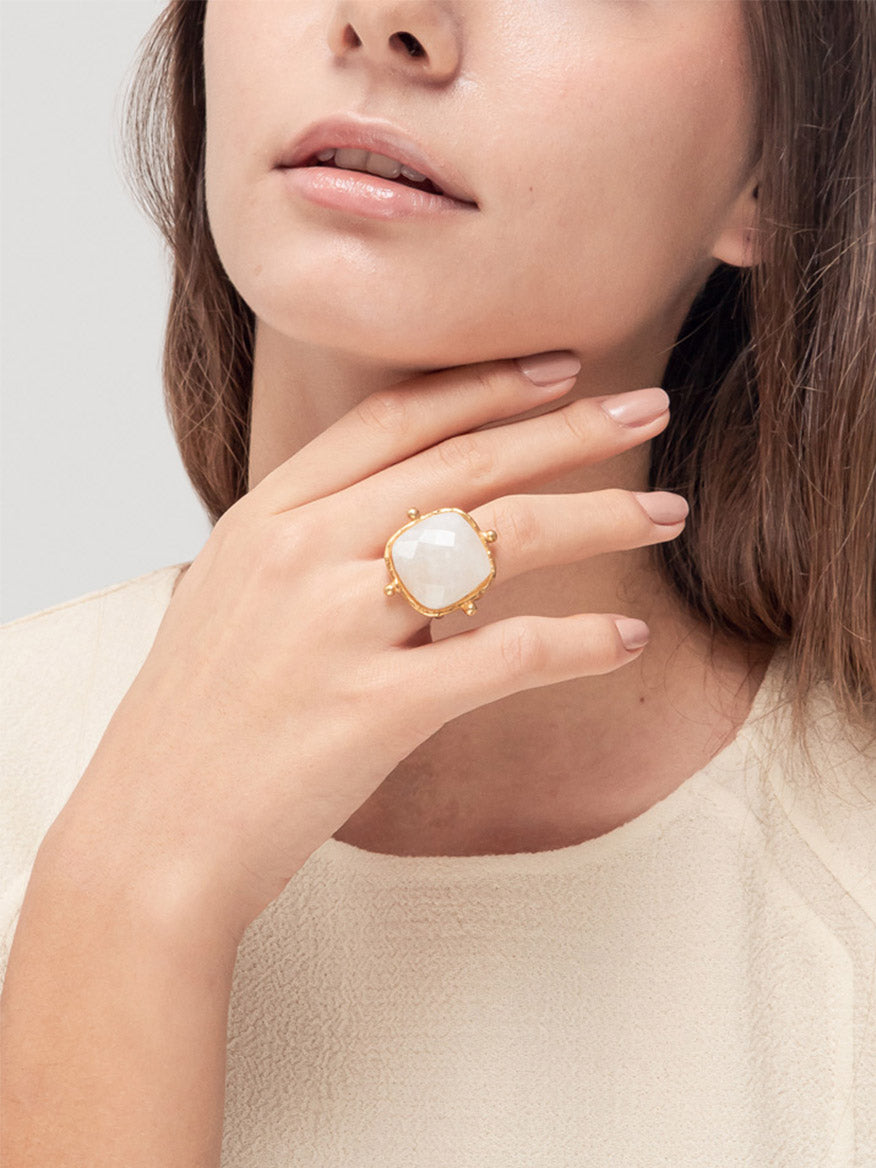 A woman wearing a cream top showcases the stunning Sylvia Toledano Ring Cushion featuring a large Moonstone, set in a square shape with a delicate gold band on her left hand, elegantly positioned near her chin.