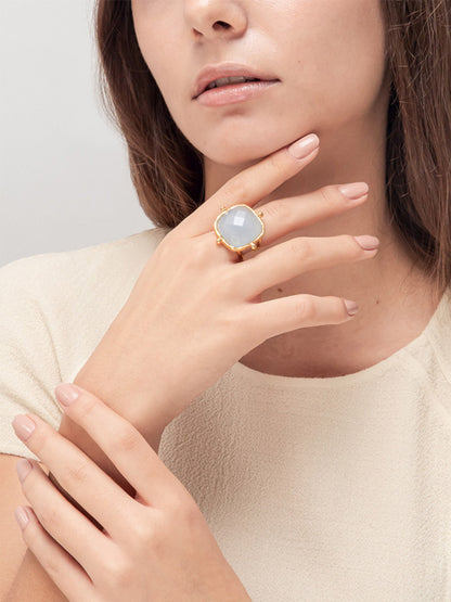 A close-up of a person wearing the exquisite Sylvia Toledano Ring Cushion in Chalcedony, with its fine gold design accented by a pale Chalcedony gemstone, as they rest their hand elegantly near their face.