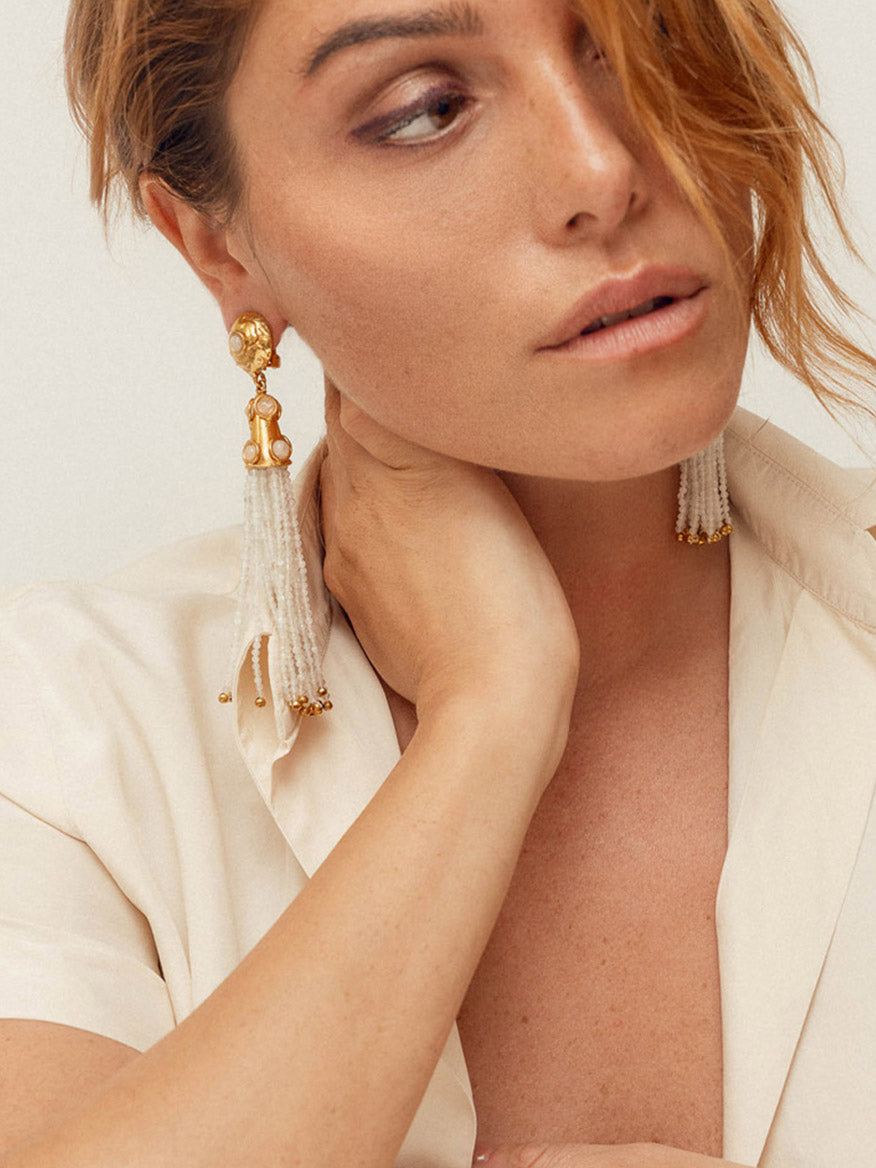 A woman with short brown hair wears a cream-colored top and Sylvia Toledano Earrings Gio in Moonstone, brass gilded with fine gold, adorned with tassels. Resting her hand at the base of her neck, she gazes slightly downward.