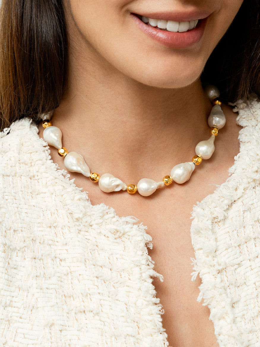 A close-up of a person wearing the Sylvia Toledano Necklace Grace in Gold & Baroque Pearls, featuring alternating white Baroque pearls and brass gilded gold beads, paired with a textured light-colored garment.