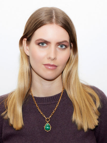 A woman with long blonde hair is adorned in a purple top, her elegance accentuated by a Sylvia Toledano Pendant Drop Gold Chain featuring a striking green malachite stone, standing gracefully against a plain background.