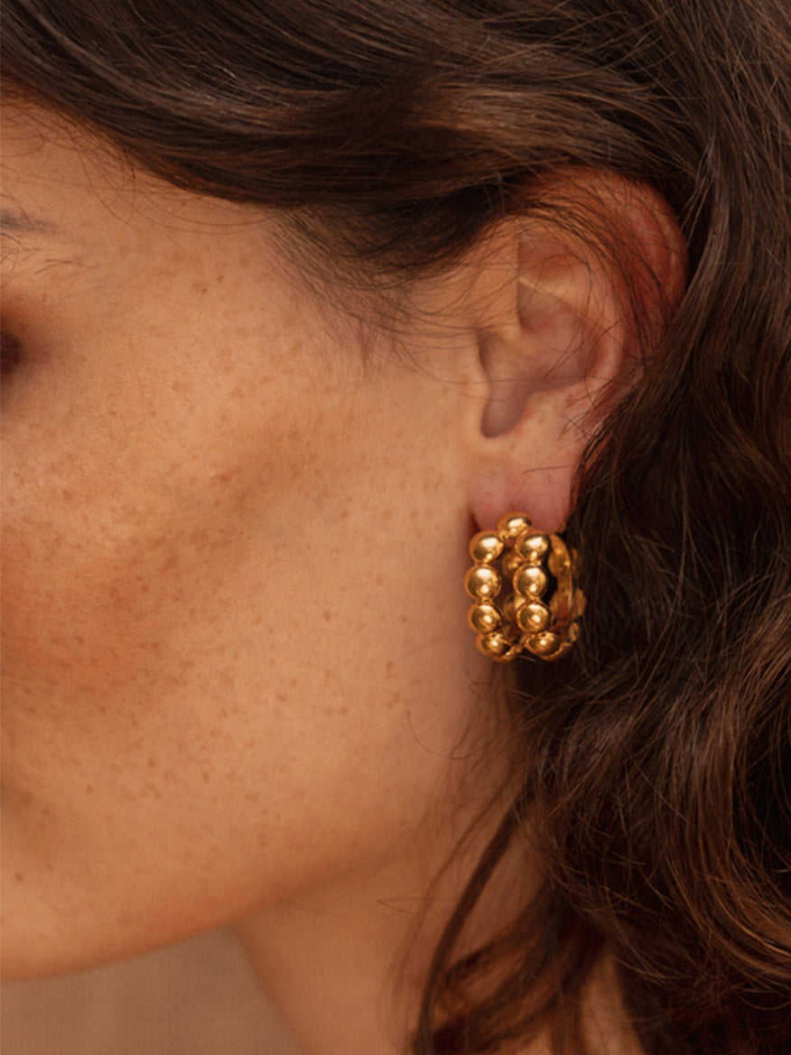 Close-up of a person's ear wearing *Sylvia Toledano Earrings Tribal in Gold* adorned with multiple small, gilded spheres. The person's dark and wavy hair partially covers the ear.