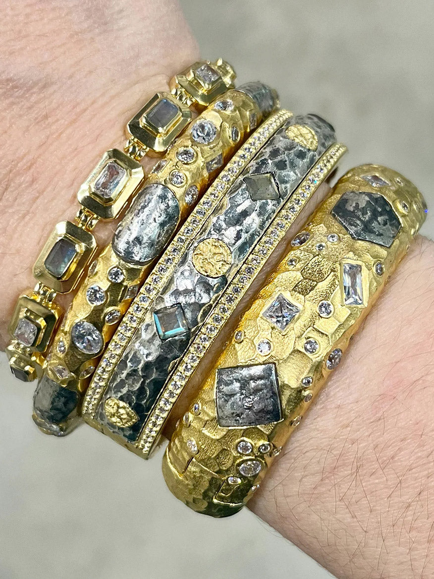 A wrist adorned with four Tat2 Designs Labradorite Mota Coin Bangles in Gold & Vintage Silver, featuring baroque pearls, variously cut gemstones, and hammered textures.