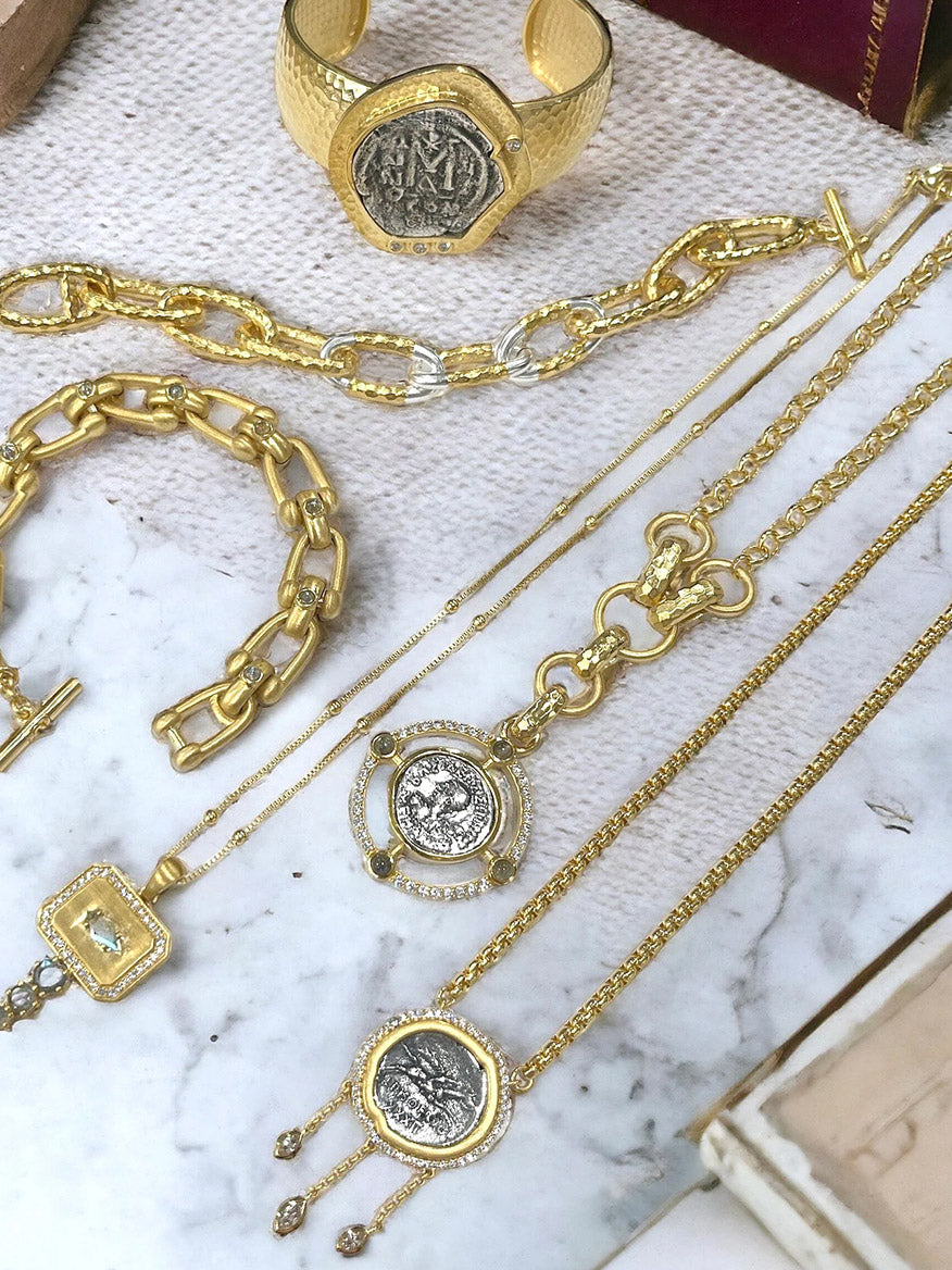 The Tat2 Designs Miko Coin Necklace in Gold & Vintage Silver, featuring coin and chain designs, is displayed on a marble and textured surface.