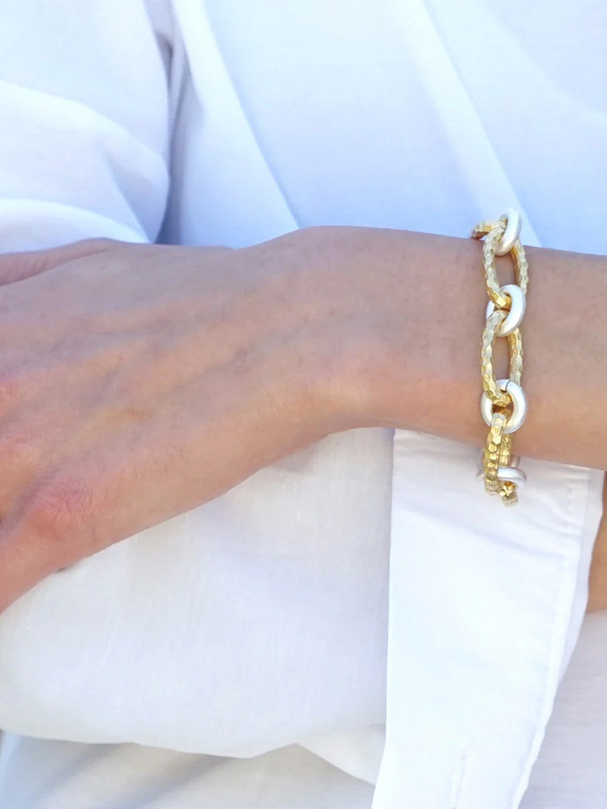 A person wearing a white shirt and the Tat2 Designs Ravelle Hammered Chain Bracelet in Gold Two-Tone, featuring custom metal textures, on their wrist.