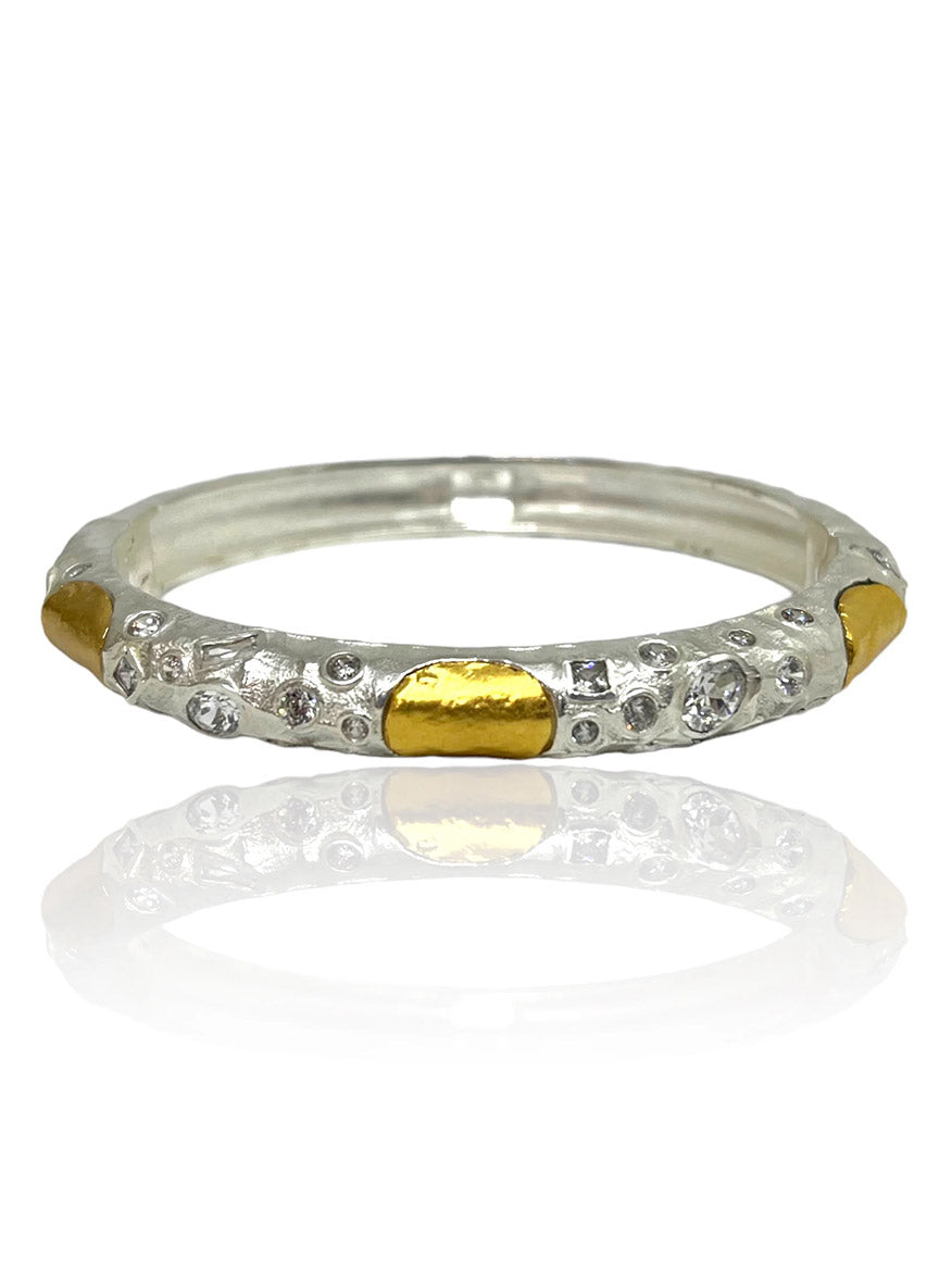 Tat2 Designs Cela Bangle in Satin Silver features textured detailing, small clear cubic zirconia stones, and gold oval accents reminiscent of gold-plated coins, evenly spaced around the bangle. It is shown on a white reflective surface.