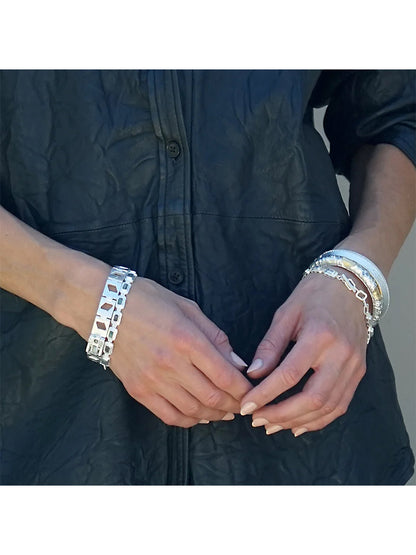 A person wears a textured black button-up shirt and multiple silver bracelets, including the Tat2 Designs Cela Bangle in Satin Silver, with their hands loosely clasped together at waist level.