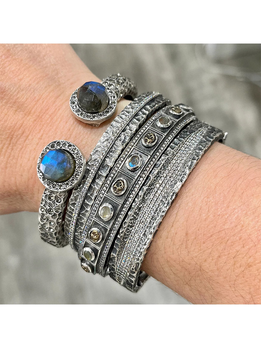 A wrist is adorned with layered silver bracelets, some featuring textured patterns and others set with round gemstones, including two prominent blue stones. Among the stack is the standout Tat2 Designs Egy Crystal Line Bangle in Vintage Silver, showcasing Cubic Zirconia accents and a sleek magnetic hinge closure for effortlessly stylish wear.