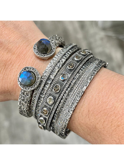 A wrist is adorned with layered silver bracelets, some featuring textured patterns and others set with round gemstones, including two prominent blue stones. Among the stack is the standout Tat2 Designs Egy Crystal Line Bangle in Vintage Silver, showcasing Cubic Zirconia accents and a sleek magnetic hinge closure for effortlessly stylish wear.