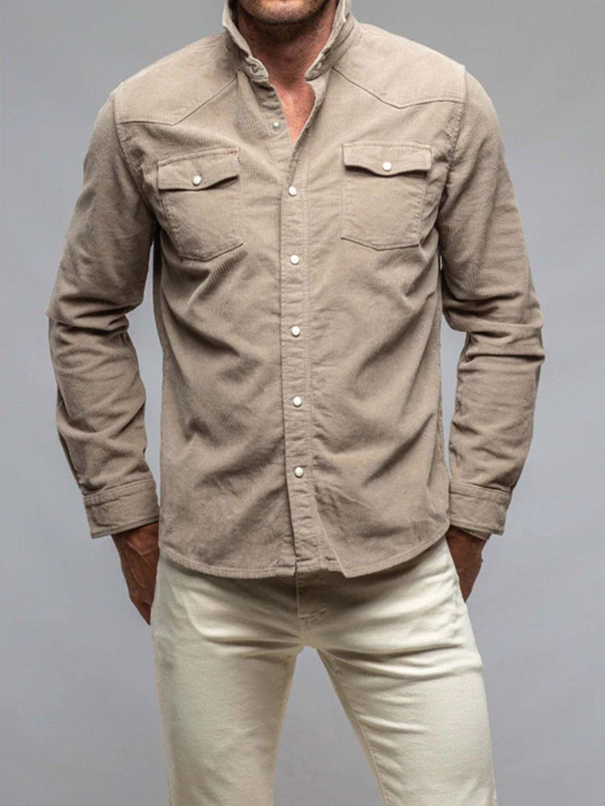 A man in a Teleria Zed Roper Western Snap Shirt in Tortora and cream pants stands against a gray background, his hands tucked casually in his pockets.