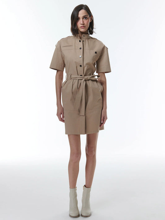 A woman posing in THEO The Label Thallo Safari Dress in Sand with a military-inspired design and white ankle boots against a plain background.
