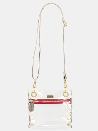 The Hammitt Los Angeles Tony Small Crossbody Bag in Clear Pewter is a stadium-approved clear bag that features a red zipper on the front, gold rivet details, and two gold rings attaching the adjustable crossbody strap. It is crafted with TPU and leather trim for added style and durability.