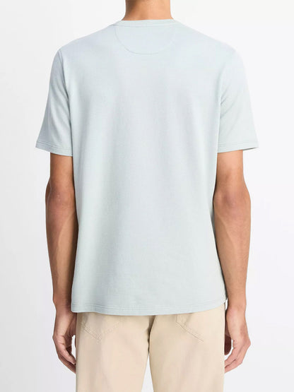 A person stands facing away, wearing the Vince Birdseye Jacquard Pima Cotton T-Shirt in Skylight and beige pants against a plain background.