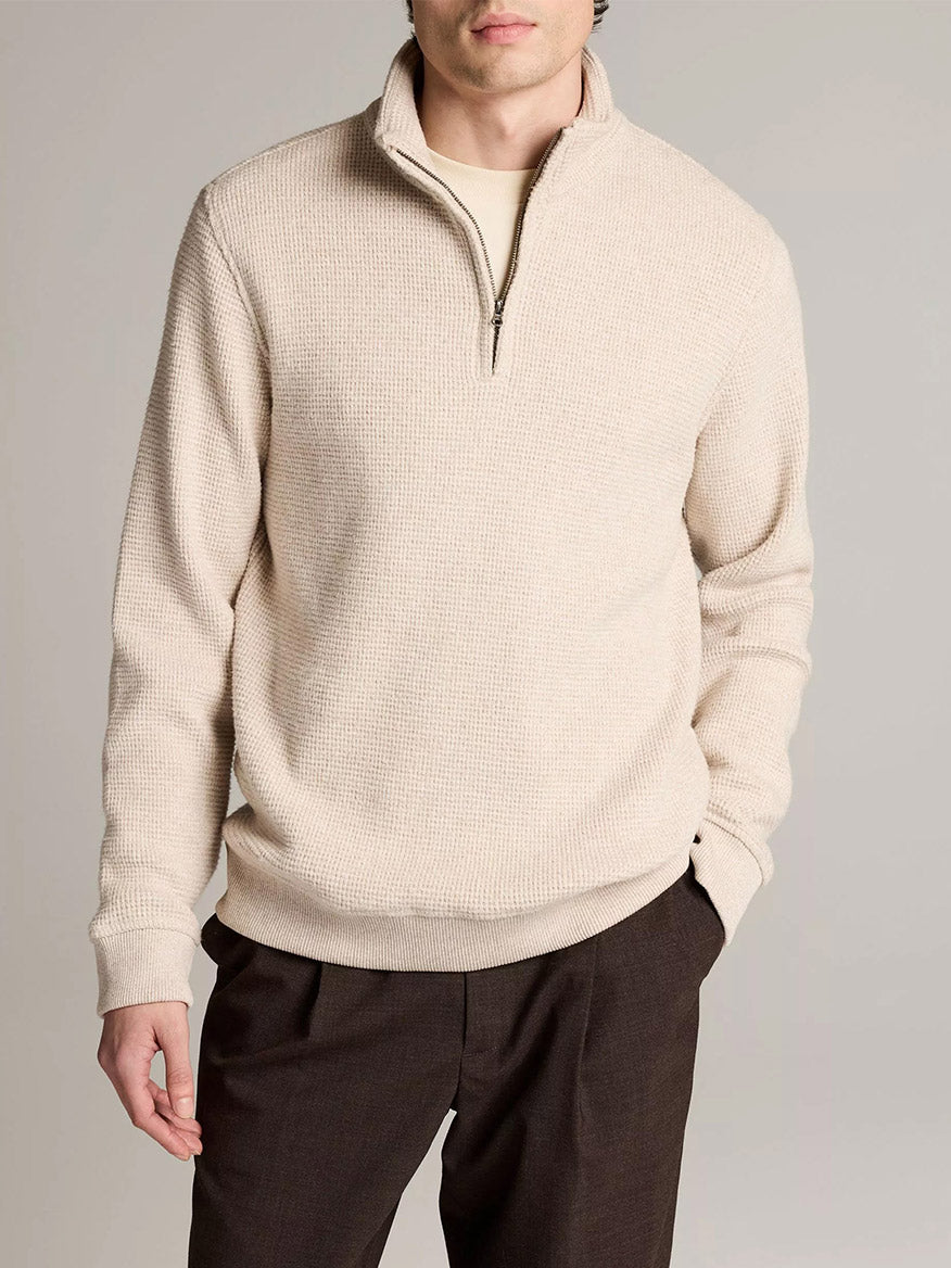 A man wears the Vince Brushed Cotton-Blend Waffle Quarter-Zip Pullover in Heather Oatmeal with dark pants, standing with one hand in his pocket against a plain background.