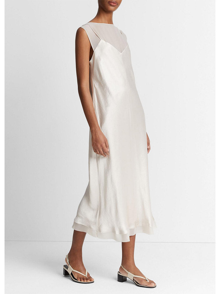 Woman in a white Vince Chiffon-Layered Satin Slip Dress in Champagne with asymmetrical neckline and black and white sandals, standing against a plain background.