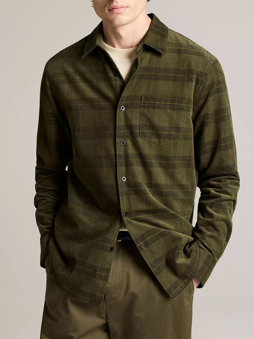 A man wears the Vince Hamilton Plaid Cotton Corduroy Button-Front Shirt in Night Leaf Combo over a beige tee with matching olive green pants, hands in his pockets.
