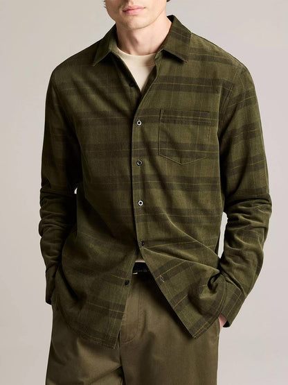 A man wears the Vince Hamilton Plaid Cotton Corduroy Button-Front Shirt in Night Leaf Combo over a beige tee with matching olive green pants, hands in his pockets.