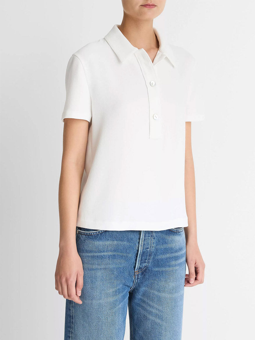 A person wearing the Vince Crepe Polo Shirt in Off-White with agoya shell buttons and blue jeans stands against a plain light background.