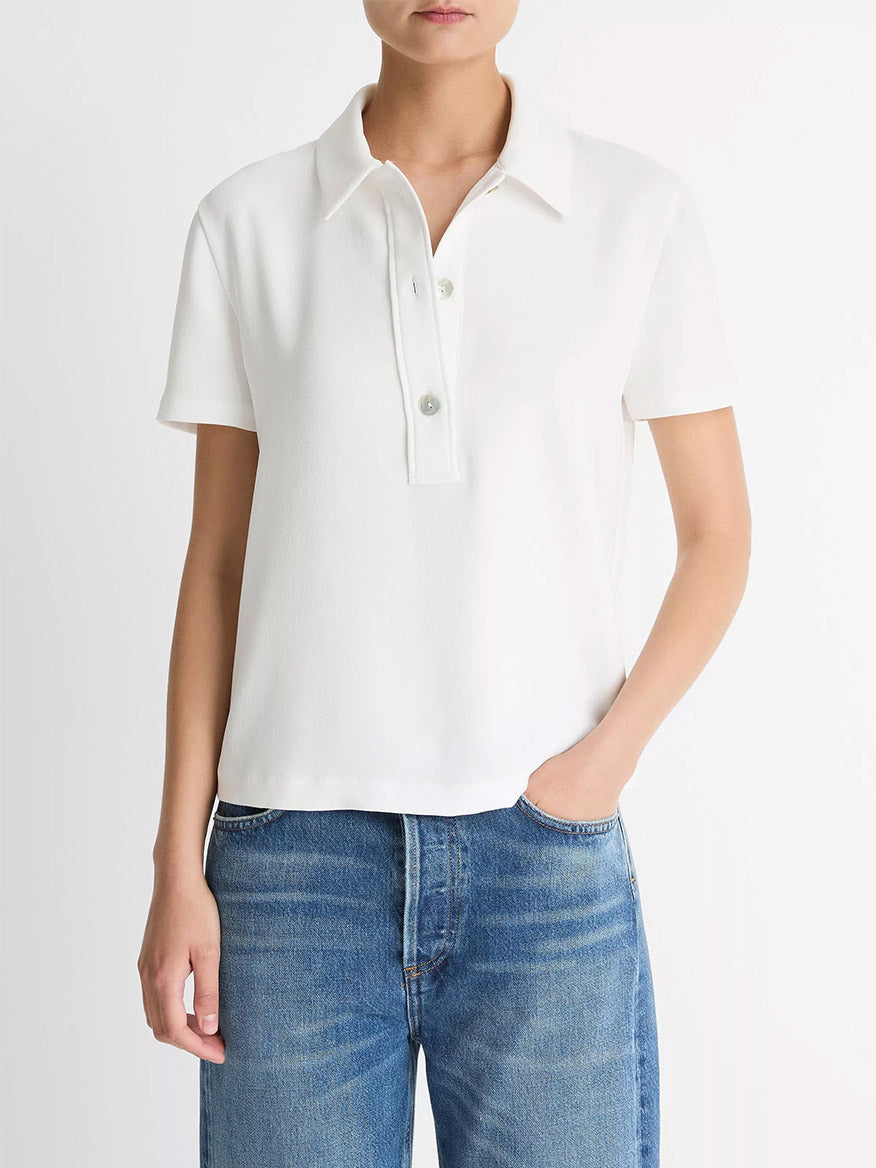 A person wears the Vince Crepe Polo Shirt in Off-White with blue jeans, featuring agoya shell buttons, standing against a plain white background.