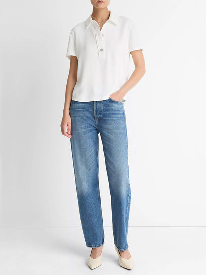 A person stands against a white wall wearing the Vince Crepe Polo Shirt in Off-White with agoya shell buttons, blue straight-leg jeans, and beige pointed-toe shoes.
