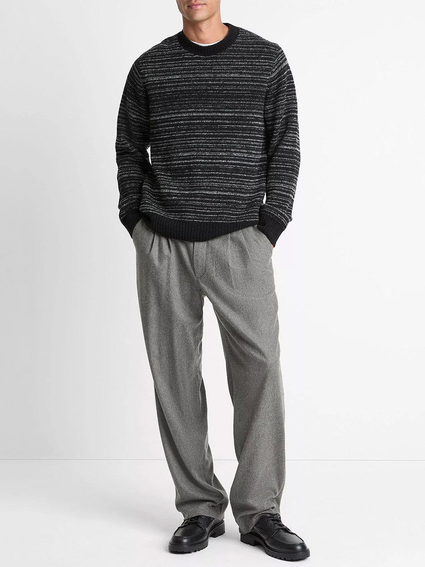 A person wearing a Vince Marled Stripe Merino Wool-Blend Sweater in Black, loose gray trousers, and black shoes stands against a plain white background.