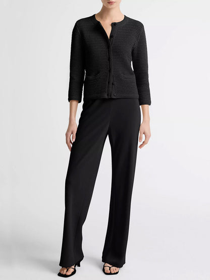 A woman stands against a plain background wearing the Vince Italian Cotton Crochet Cardigan in Black, styled with black wide-leg pants and black heeled sandals—ideal for spring-summer layering.