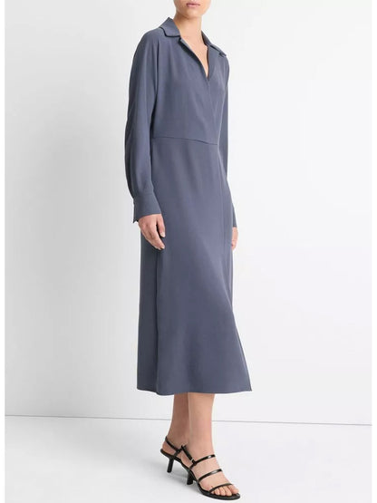 A person is wearing the Vince Shaped Collar Dolman-Sleeve Dress in Dark Water, crafted from fluid crepe, standing against a plain white background. The dress features a V-neck and falls to mid-calf. They are also wearing black heeled sandals.