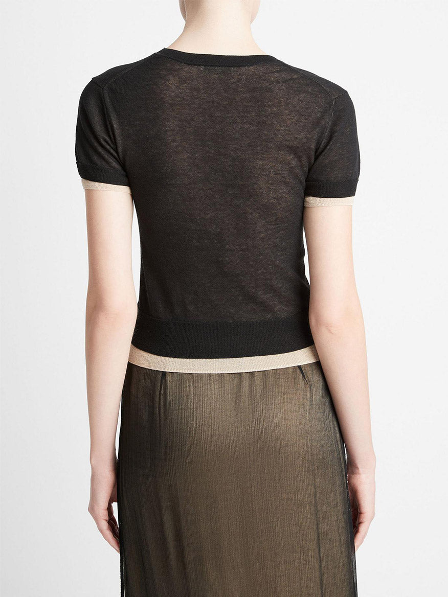 Rear view of a woman wearing a Vince Double Layer Knit T-Shirt in Black/Oat Sand Combo and a metallic pleated skirt, standing against a neutral background.