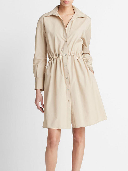 A woman in a Vince Cotton Drawcord Ruched Shirt Dress in White Oak with long sleeves, drawcord waist, and buttons down the front, stands facing left with her face not visible.