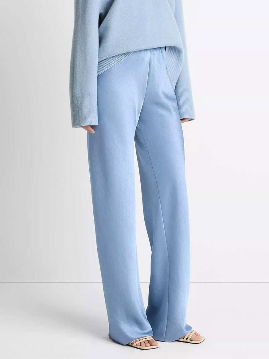 A person wears the Vince Fluid Satin Bias Pant in Pacific with a matching sweater and open-toe sandals, standing against a plain background.